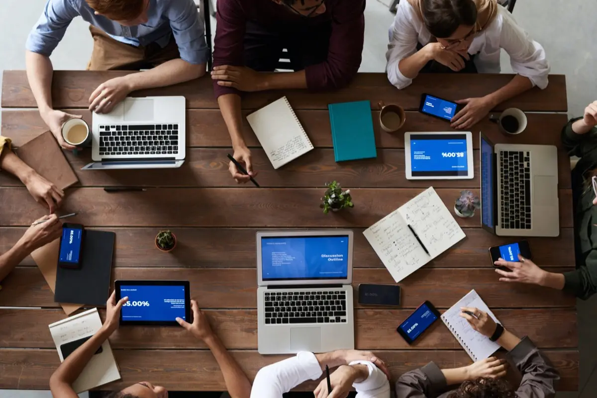 Vista de cima de uma mesa de reunião com sete pessoas em volta, notebooks, tablets mostrando gráficos e cadernos de anotação.